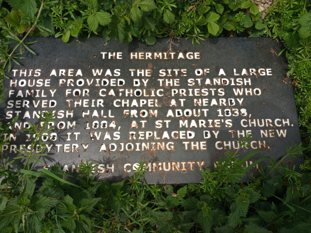 Dark gray, rectangular metal plaque, deeply weathered and showing signs of rust and age. The plaque is embedded in the earth, partially surrounded by lush green vegetation. The plaque's surface bears an inscription in white, slightly faded lettering, detailing the history of a hermitage. The text is in a serif font, characteristic of old-fashioned signage. There are minor irregularities in the lettering, possibly due to age and weathering. The plaque's position is slightly angled, not perfectly flat against the ground. The plaque is nestled within a dense bed of various green plants. These include small, rounded leaves reminiscent of ground cover, taller, slender stems with small, pointed leaves possibly of a weed variety, and some larger, more textured leaves suggesting stinging nettles. The ground itself is not directly visible, being completely covered by the vegetation. 