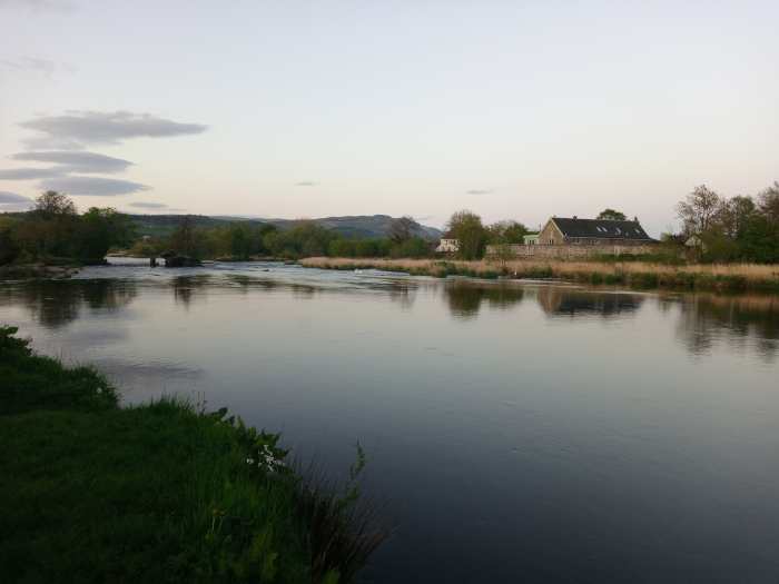 Tranquil river scene. The river, calm and reflective, dominates the foreground and middle ground, its dark, still water mirroring the sky and surrounding landscape. A stone building, appearing to be a large house or possibly a converted barn, sits on the right bank of the river in the mid-ground. It's a low, long structure with a dark roof and light-colored stone walls, showing evenly spaced windows. No people or animals are visible. The left bank of the river features a line of lush, dark-green vegetation that grows down to the water's edge. Slightly further back, on the left side, there is what appears to be a low-lying weir or dam structure where the river's current seems to slightly break.