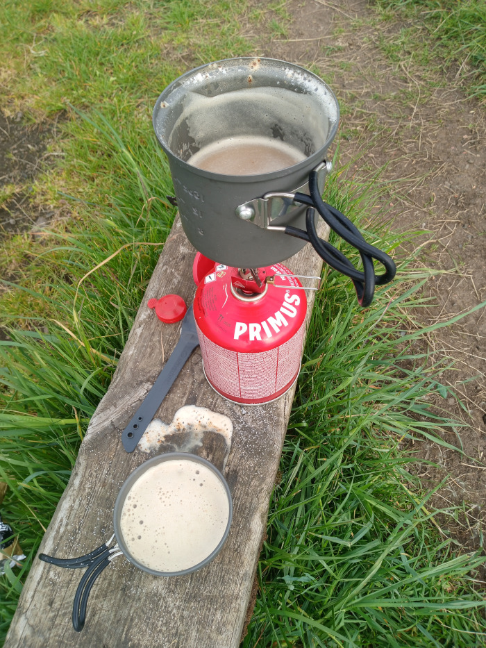 Rustic outdoor scene depicting the preparation of a hot beverage, likely coffee or tea, using a portable camping stove. Dominating the foreground is a weathered, gray-brown wooden bench or log. Resting on it are two dark gray metal camping mugs. The top mug, larger, sits atop a Primus brand red canister stove; it's filled with a light brown, frothy liquid, suggesting recently boiled or brewed beverage. The second mug, smaller, sits below and to the right, also containing a similar light brown, frothy liquid.  A dark gray, spatula-like utensil lies next to the stove and a small red cap is near the stove's base.