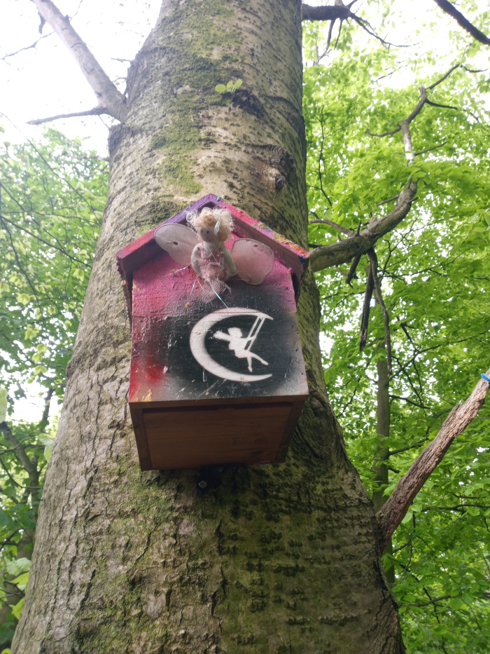 Small, handcrafted birdhouse-like structure affixed to the trunk of a large tree. It's painted in a vibrant, graffiti-style; a pinkish-red base overlaid with darker shades creating a mottled effect, accented by a stark black crescent moon symbol incorporating a silhouetted fairy figure swinging on a swing. Atop the house sits a small, handcrafted fairy figurine with delicate wings, seemingly poised in a protective stance. The fairy is light-coloured with a subtle, rustic look, almost ethereal against the bold colours of the house.