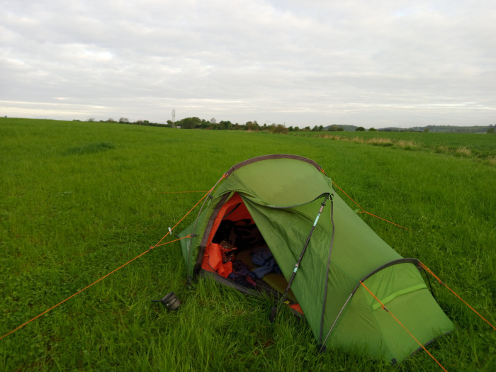 Vibrant green, two-person backpacking tent pitched in a lush green field. The tent is a single-wall design, with orange accents visible along the seams and guy lines. Its vestibule is partially visible, and the interior appears to be partially set up with sleeping bags or other camping gear. The interior is mostly dark, making details indistinct. A small, dark-colored object (possibly a camping mug or other small item) lies on the grass near the tent's base. The tent's poles are visible, showing it to be a lightweight and easily assembled design. No people are present. The grass is short, uniformly colored, and appears soft and slightly damp. In the background, a slightly darker green line indicates the horizon, barely separating the field from the sky. The sky is a pale, overcast gray-white, devoid of strong sunlight. The overall lighting is soft and diffuse, suggesting either early morning or late afternoon light. The dominant colours are the bright green of the field and the olive-green of the tent, contrasted by the subtle orange of the tent's details.