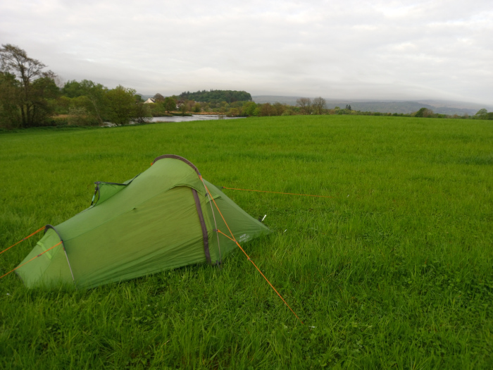 Vibrant green, single-person backpacking tent, pitched in a sprawling field. It's a dome-style tent with a slightly asymmetrical shape, its flysheet taut but not fully extended, suggesting it's either recently pitched or being packed up. The tent's fabric appears subtly textured, with hints of sheen reflecting the muted light. Orange guy lines radiate from the tent's corners and stakes, barely visible in the grass. There are no people present. The tent sits in a vast, flat field of lush, bright green grass, which stretches to the horizon. The grass is unbroken, save for a few small, scattered wildflowers. In the distance, a low-lying, gently undulating landscape is visible. It's a mixture of muted greens and browns from distant trees and possibly hills, suggesting a rural or pastoral setting. A calm body of water, possibly a river or canal, is faintly visible on the horizon, providing a horizontal line that accentuates the flatness of the field. The sky is an overcast, pale grey-white, casting a diffuse, soft light across the scene; there are no harsh shadows.