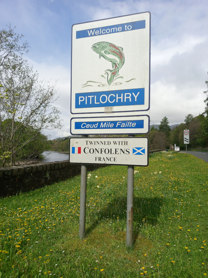 Vertically stacked, rectangular road signs standing proudly in a vibrant meadow. A bright white sign with bold blue lettering proclaiming Welcome to PITLOCHRY in a sans-serif font. A stylised illustration of a salmon leaping from water occupies the top portion. The style is slightly cartoonish, yet detailed enough to show scales and fins. The salmon is a muted green and red. A smaller white sign beneath the main one, displays Ceud Mile Failte in a smaller, similar font. This is Gaelic for A Hundred Thousand Welcomes. The smallest sign, this one features Twinned with CONFOLENS FRANCE in crisp, clear lettering. Small French and Scottish flags flank the town's name. The overall design is simple and clean.