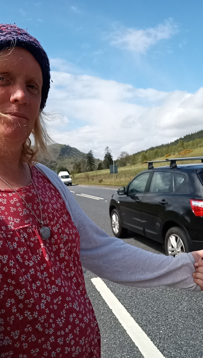 Fair-skinned person with shoulder-length blonde hair partially obscured by a deep purple and blue knitted beanie. They are wearing a red dress adorned with a small white floral print, and a light grey, long-sleeved cardigan. A silver necklace with a dark grey or black pendant hangs around their neck. Their arm is outstretched towards the viewer's left, as if hitch-hiking or gesturing for a vehicle to stop. Their expression is neutral, perhaps slightly pensive. Their hand is slightly blurred, suggesting motion. In the background, a black SUV with roof racks is visible, stopped or slowing down on a highway. A smaller, light-colored car is also faintly visible further back. The setting is a brightly lit highway, bisected by a double white line, under a mostly clear blue sky. Fluffy white clouds are scattered across the upper portion of the frame. In the far background, a soft green landscape of rolling hills dotted with sparse trees is visible. The lighting suggests daytime, with the sun casting even illumination on the scene. The color palette is naturalistic, with the cool blues of the sky contrasting against the warm reds and greens of the clothing and landscape. The highway itself is dark grey asphalt.