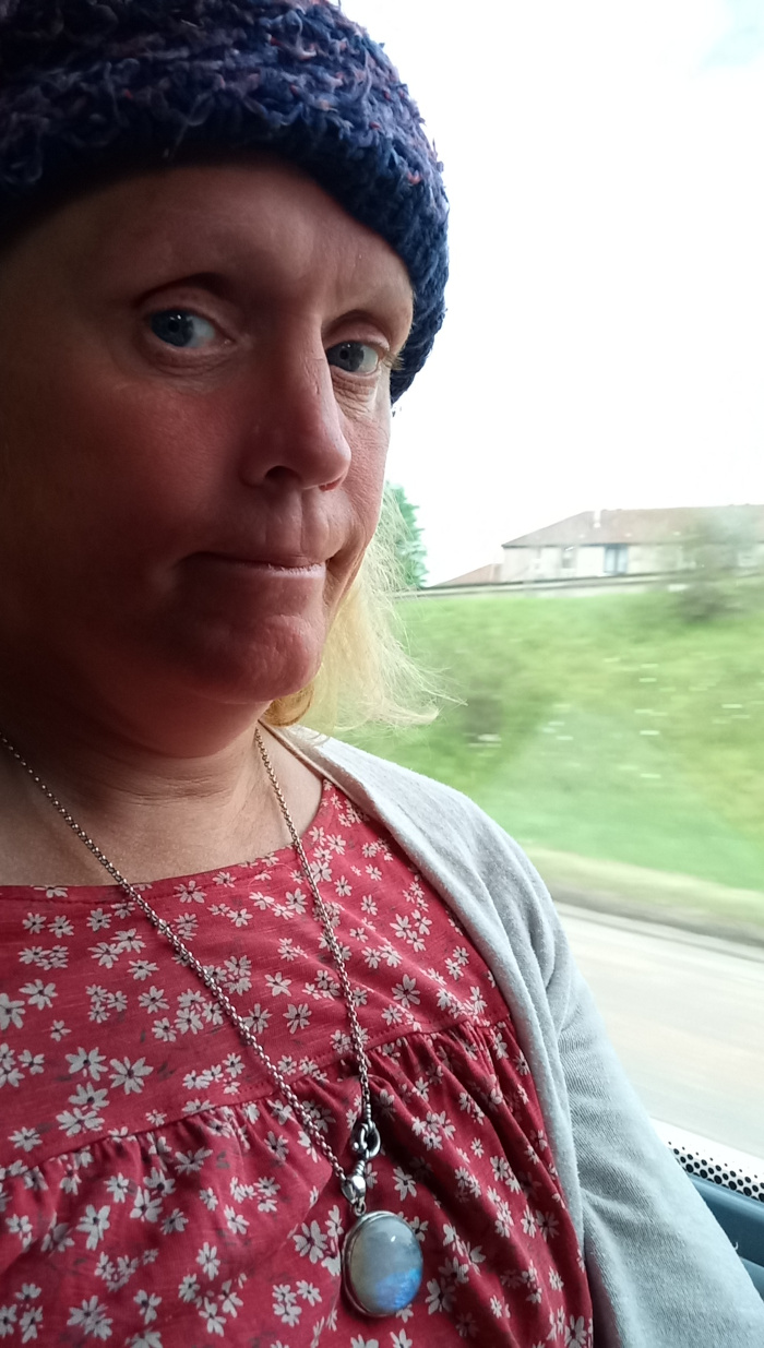 Close-up portrait of a fair-skinned woman, possibly middle-aged, with blonde hair partially visible beneath a deep purple knitted beanie. The beanie is slightly askew, revealing her forehead and temples. Her expression is subtly melancholic; her mouth is slightly downturned, and her eyes, although clear and directly engaging the camera, hold a pensive quality. She's wearing a red dress with a small, white floral pattern, and a delicate silver chain necklace featuring a round moonstone pendant. The pendant is smooth and reflects a soft, ethereal light. She is seated, her upper body angled slightly towards the camera, and her gaze is directed slightly to the right. Her hands are not visible. A grey cardigan or jacket is draped over her shoulders. The background is blurred, suggesting a shallow depth of field, indicative of a photograph taken through a vehicle window. The focus is strongly on the woman's face and upper body. The out-of-focus background features a verdant landscape with rolling hills and a low building in the distance.  The lighting appears natural, likely daylight, casting a soft glow on the woman's face, slightly highlighting the textures of her skin and the beanie's yarn. The color palette is relatively muted: deep purple in the beanie, muted reds and whites in the floral dress, soft grey in the cardigan, and the contrasting green hues of the blurred background. The moonstone pendant adds a cool, silvery tone.