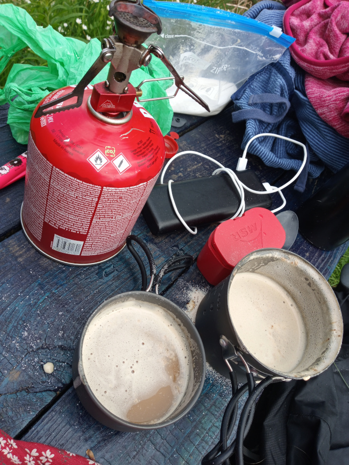 Camping scene, specifically the preparation of hot drinks. A red, cylindrical propane canister (likely for a portable stove) is prominently placed on a dark blue wooden table. It's covered in small print detailing safety warnings and product information. Atop the canister sits a small, folded, silver-coloured portable camping stove. Two grey metallic camping pots, nested slightly, sit lower in the frame. Both pots contain a light beige, foamy liquid, likely a hot beverage. The pots are connected by a black cord with a simple handle. Next to the pots, there's a small scattering of a light-coloured powder, possibly powdered milk or another ingredient for the drink. A red, heart-shaped, compact power bank is also present on the table. Other items strewn around include: a partially visible transparent plastic bag with white powder inside (possibly powdered milk or sugar), a dark blue sweatshirt and a pink fleece partially rolled up, a white charging cable connected to a black power bank (likely for a phone), a partially visible pink marker, and a crumpled green plastic bag. A black backpack is visible in the bottom right corner. 
