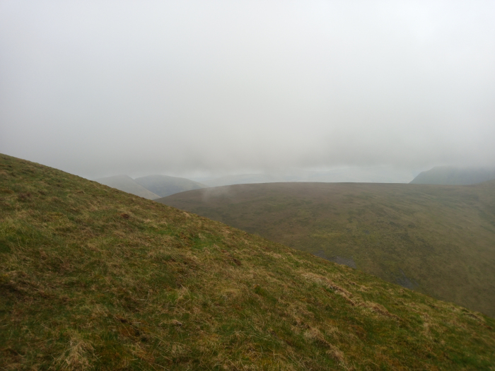 Gently sloping hillside covered in sparse, brownish-green grass. This hillside occupies the majority of the foreground and extends to the mid-ground, where it merges into rolling hills shrouded in fog. The scene is a high-altitude landscape, likely a mountain range, dominated by a thick, pervasive fog or low-lying clouds. The sky is a uniform, light gray, indistinguishable from the fog. The fog obscures the details of the distant hills, leaving them as soft, hazy shapes. The overall colour palette is muted and subdued, consisting primarily of various shades of gray and brownish-green. The grass on the hillside is a mix of dull greens and browns, suggesting either late autumn or early spring. The lighting is diffuse and soft, lacking strong shadows due to the overcast conditions.
