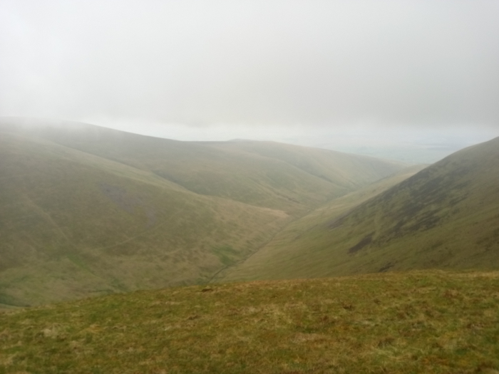 high-altitude landscape, likely a mountain range or moorland. The foreground is a gently sloping hill covered in short, light brown-green grass, suggesting a relatively sparse, possibly windswept, vegetation. The mid-ground and background are dominated by a series of rolling hills and valleys extending into the distance. A valley cuts through the middle of the image, receding into the mist. The overall colour palette is muted, dominated by various shades of light brown, green, and grey. The lighting is diffuse and soft, characteristic of an overcast day. The fog or mist hangs low, obscuring the far distance and giving a hazy, indistinct quality to the scene. The light is not harsh or dramatic; rather, it's a muted and even light that minimises shadows. The composition is panoramic, taking in a wide expanse of the landscape. The perspective is from a high vantage point, looking down and across the valleys and hills. The viewpoint seems to be located on a slightly elevated position within the foreground hills, providing a fairly broad sweep of the terrain. The leading lines of the hills draw the viewer’s eye towards the valley and the receding distance.