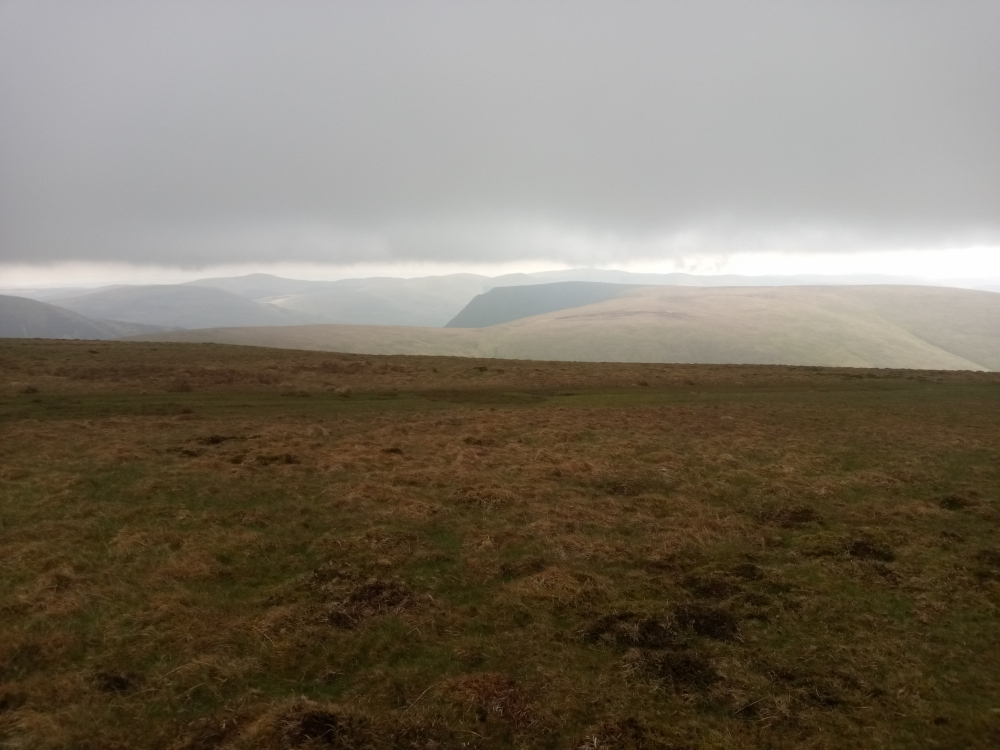 Vast, open expanse of upland moorland stretching from the foreground to the middle ground. The foreground is dominated by a field of low-lying, dry, brownish-green vegetation, appearing somewhat sparse and windswept. Patches of darker brown indicate areas of more compacted earth or perhaps subtly different plant life. There are no people or animals visible. The background consists of rolling hills and mountains under a heavy, overcast sky. The mountains are muted in colour, shades of grey-green and brown blending seamlessly into the pale grey of the sky. The sky occupies a significant portion of the upper two-thirds of the image, forming a massive, diffuse light source, casting a soft, even light across the landscape. There's no harsh shadowing; the lighting is flat and diffused, creating a mood of muted tranquillity, perhaps hinting at an impending rain or recent precipitation.