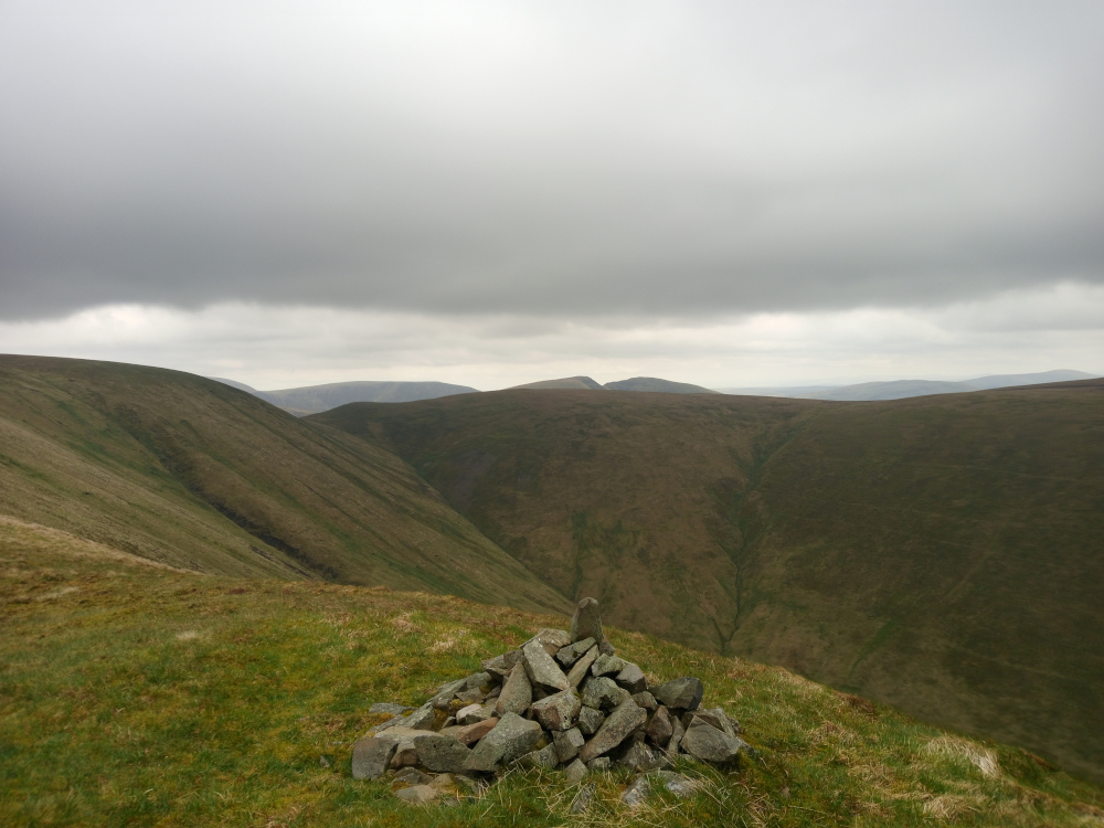 Modest cairn, a small, meticulously stacked pile of grey and brown rocks, situated on a gently sloping hilltop. The rocks vary in size and shape, with some larger, angular stones forming the base and smaller, flatter stones creating a slightly peaked apex. There is no human presence. The scene is a vast, undulating landscape of rolling hills and valleys under a heavy, overcast sky. The hills are a muted palette of greens and browns, the grass showing hints of yellowish-brown in places, suggesting either dry conditions or early spring growth. The valleys are darker, creating a strong contrast with the lighter slopes. The sky dominates the upper two-thirds of the image, a thick, uniform layer of grey cloud with minimal variation in tone, suggesting a moody, possibly slightly threatening weather. The light is diffuse and soft, lacking strong shadows, characteristic of an overcast day.