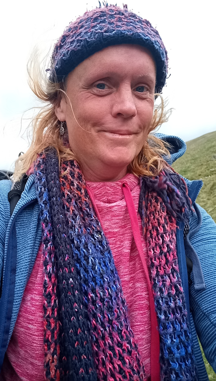 Selfie portrait of a fair-skinned woman with windblown, shoulder-length blonde hair. She is positioned slightly off-center, looking directly at the camera. Her expression is a soft, almost subtle smile, conveying a sense of calm contentment. She is wearing a snug-fitting, dark blue ribbed jacket over a vibrant pink heather-toned hoodie. A chunky, hand-knitted scarf, predominantly dark navy blue with splashes of deep red, pink, and blue, is loosely draped around her neck. A matching knitted beanie cap is pulled down to her eyebrows, enhancing her features and offering a contrast to her blonde hair. Small, silver dangling earrings are visible in her ears. A portion of a dark blue backpack strap is visible on her left shoulder.