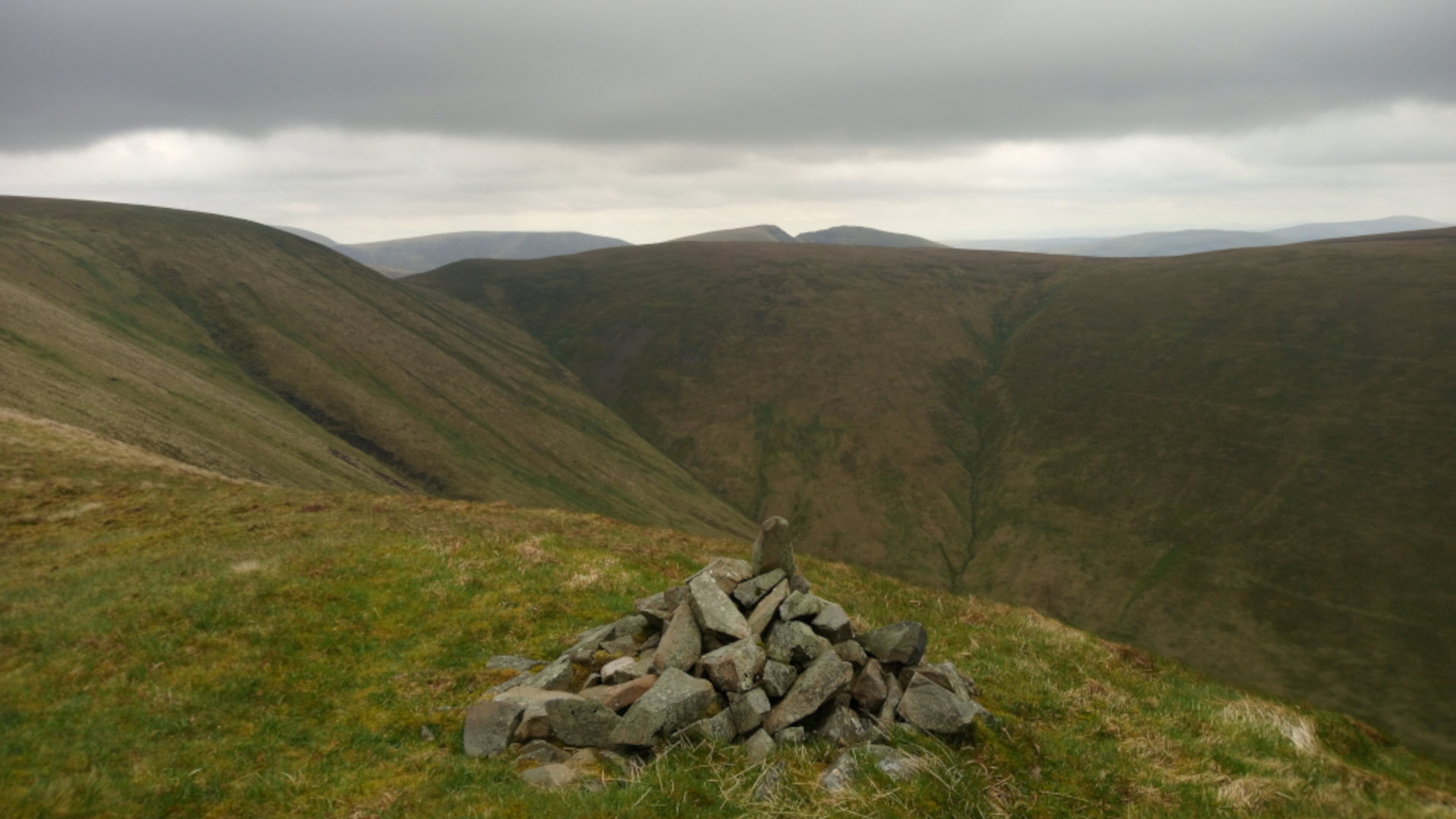 Sweeping expanse of rolling, heather-covered hills under a brooding, overcast sky. The foreground consists of short, slightly mossy grass, a pale yellowish-green in hue. The hills in the mid-ground and background are a deeper, richer green, tinged with brown in the more distant areas. The color palette is muted and earthy, dominated by various shades of green and brown, with the sky a dull, greyish-white. The lighting is diffuse and soft, characteristic of an overcast day; there are no harsh shadows, creating a somewhat sombre and subdued mood. The valleys between the hills show muted shades of brown and green and are partially obscured by the distance and the atmospheric perspective.