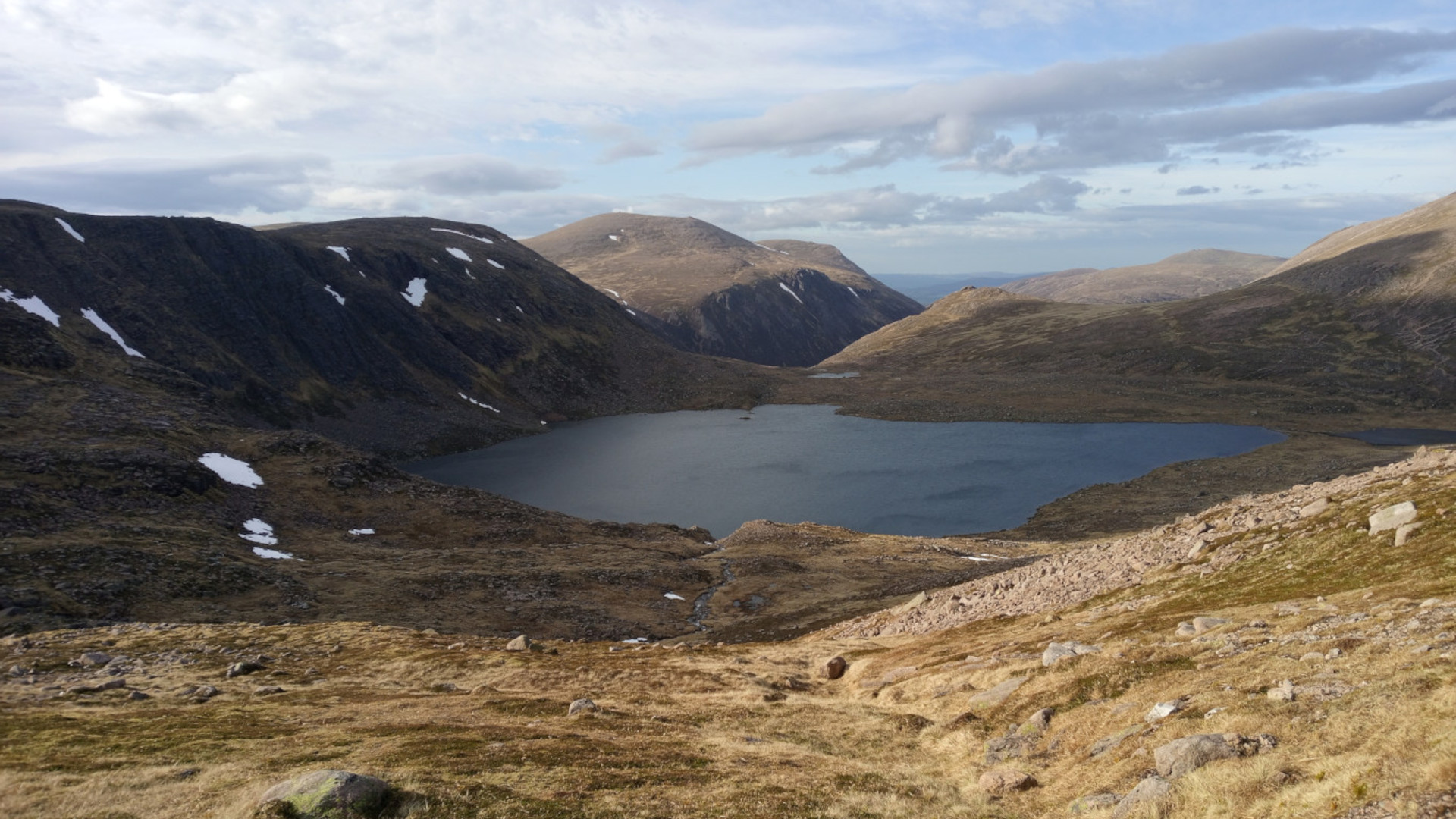 A high-angle, long shot of a serene mountain lake nestled within a rugged, mountainous landscape. The lake, dark and still, is surrounded by steep, rocky hillsides sparsely covered with low-lying vegetation. Patches of snow are visible on the higher slopes, suggesting a cold or high-altitude environment. The sky is partly cloudy, with a mix of blue and white, adding depth and texture to the scene. The overall impression is one of wild, untouched beauty.