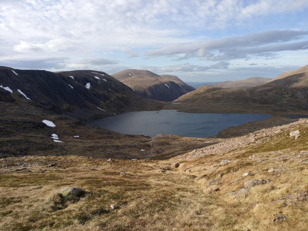 High-angle, long shot of a serene mountain lake nestled within a rugged, mountainous landscape. The lake, dark and still, is surrounded by steep, rocky hillsides sparsely covered with low-lying vegetation. Patches of snow are visible on the higher slopes, suggesting a cold or high-altitude environment. The sky is partly cloudy, with a mix of blue and white, adding depth and texture to the scene. The overall impression is one of wild, untouched beauty.