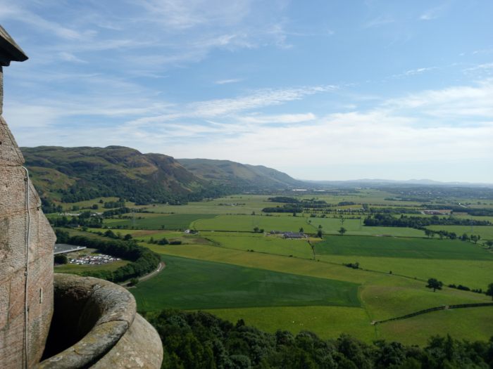 Prominent, weathered stone structure in the lower left foreground. It appears to be part of a wall or tower, possibly from a castle or historical building, with a visible, cylindrical, hollowed-out section suggesting a crenel or embrasure. The stone is a muted greyish-brown, showing signs of age and weathering. There are no people visible in the image. The scene is a panoramic view from an elevated vantage point, overlooking a vast expanse of rolling, verdant farmland. The land stretches to the horizon, showing a patchwork of varying shades of green fields, indicating different crops or grazing areas. In the mid-ground, low-lying hills are visible, their slopes covered with lush green vegetation, suggesting pasture or forest. The hills rise gently towards a range of higher, more rugged, dark-green mountains in the background. The mountains appear hazy and distant, creating a sense of depth and scale. The sky is a brilliant, clear light blue, with a few wispy, white clouds scattered across it. The lighting suggests it’s likely midday, with bright, even sunlight illuminating the landscape. The overall color palette is naturally warm, with various shades of green dominating the landscape, complemented by the earthy tones of the stone structure and the clear blue of the sky.