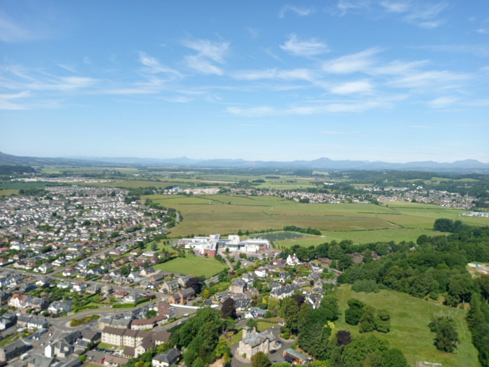 Sprawling, verdant pasture dominating the foreground. The grass is a vibrant, deep green, short to medium length, showing subtle undulations in its surface, suggesting a gentle slope.  A dark brown, almost black, earthy path is barely visible in the very bottom left corner.  In the middle ground, a dense line of deciduous trees forms a dark, textured band against the lighter green of the pasture and the rolling hills in the background. The trees are predominantly dark green, with variations in shade suggesting depth and individual trees within the mass. The background is composed of gently rolling hills, a distant, hazy landscape of pale greens and browns, culminating in a prominent hill or low mountain that's somewhat darker in tone. There are no people or animals visible in the scene.