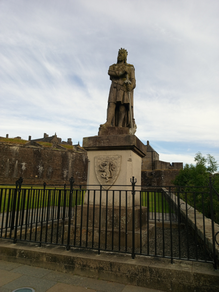 Weathered, grey-stone statue of a king, positioned atop a similarly coloured stone pedestal. The king is depicted in regal attire, wearing a crown and long robes, his arms crossed across his chest. He holds a sword at his side, partially obscured by his robes. The statue shows signs of age and weathering, with visible texture and discolouration. The pedestal features a carved stone shield bearing a heraldic emblem – what appears to be a rampant lion. The entire monument is surrounded by a simple, dark-grey metal fence, creating a protective barrier. The scene is outdoors, under a mostly cloudy sky showing soft, pale grey and white hues. The lighting is soft and diffuse, indicating an overcast day. There is a background of a large, aged stone castle wall, showing a muted range of greys and browns, indicative of aged stone. The castle shows visible stonework, some areas appearing more worn than others. Beyond the castle wall, there’s a hint of green vegetation—a grassy area in front of the monument and some trees slightly off to the right.