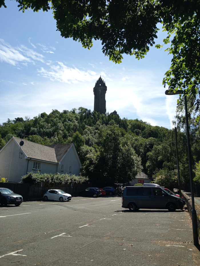 The scene is set outdoors on a bright, sunny day. The sky is a clear, light blue with a few wispy clouds. The background dominates with a lush green hill covered in dense trees, rising up to a prominent, dark gray stone tower (The Wallace Monument) that is situated centrally in the background. The tower's sharp, Gothic-inspired architecture contrasts with the soft roundness of the hill. The foreground is a paved parking area with visible parking space lines. The colours are natural and vivid: bright sky blue, deep green foliage, and the muted grays of the cars and stone monument. The lighting is strong, natural daylight casting sharp shadows. The overall colour palette is a balance between the brightness of the sky and the cool greens and grays of the landscape and architecture. The sunlight creates distinct shadows under the cars and trees. Overhanging the scene in the upper left is the shadowing foliage of a large tree. A lamppost stands prominently in the upper right background.