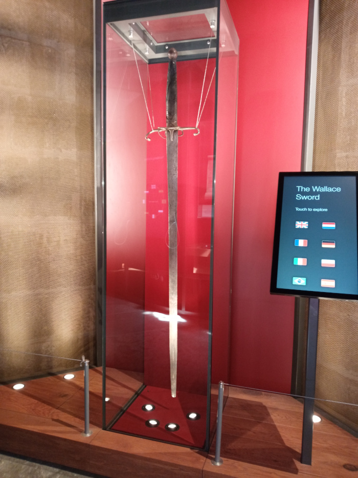 Large, ancient-looking sword, seemingly made of aged steel with a dark gray-brown patina. It's displayed vertically within a tall, rectangular, clear glass case. The sword features a simple, slightly bulbous pommel at the hilt and a relatively straight, long blade showing signs of wear and possibly damage along its length. A simple, cross-shaped guard is subtly visible near the hilt. Thin, almost invisible wires suspend the sword within the case, preventing it from touching the sides. To the right of the display case stands a sleek, black, interactive touchscreen kiosk. Its screen displays the text The Wallace Sword in a clean, sans-serif font, followed by Touch to explore. Below the text are small flag icons representing several countries (UK, Netherlands, France, Germany, Italy, possibly others). The background is primarily a rich, deep crimson red wall, providing a stark contrast to the sword and the dark glass case. The scene is set in what appears to be a modern museum setting. The floor is a dark reddish-brown wood. Subtle, recessed LED lighting is visible beneath the glass case, casting a soft glow on the base and enhancing the sword's presence. The lighting appears cool and even, minimising shadows.  The wall adjacent to the display case is textured, resembling a woven or pressed metallic material in a muted gold or bronze tone. This textural contrast adds depth to the scene.