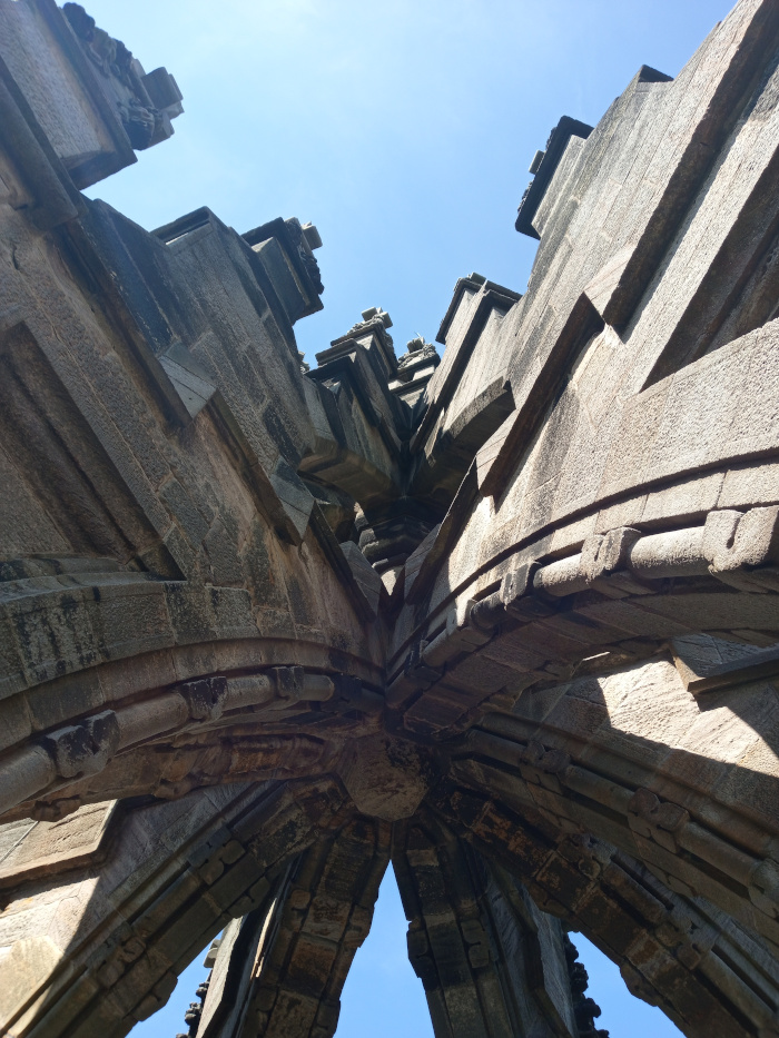 Complex, intricate stone structure, possibly part of a monument or ruin. The stonework is the dominant feature, showcasing a series of intersecting arches, buttresses, and angular blocks of varying sizes. The stone appears aged and weathered, a dark gray-brown colour with variations in tone and texture from sun exposure and age. The blocks are meticulously fitted together, creating a visually captivating design. There are no people or other objects present besides the stone structure itself. The scene is outdoors under a bright, clear blue sky. The sky acts as a stark contrast to the dark stone structure, emphasising its form and texture. The sunlight casts strong shadows within the crevices and indentations of the stone, accentuating its three-dimensional qualities and depth. The image utilises a dramatic low-angle perspective, looking directly upward into the heart of the structure. This perspective creates a feeling of grandeur and scale. The viewer's eye is drawn upwards along the converging lines of the arches and stone blocks toward the sky, creating a sense of depth and height.
