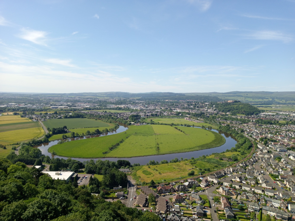 High-altitude, long shot showcasing a meandering river curving through a verdant landscape. The river, a dark, almost charcoal grey-brown, acts as a central, organic line, snaking its way through fields of vibrant, emerald green. These fields are interspersed with patches of pale yellow, suggesting agricultural activity. On the right bank of the river, a suburban residential area unfolds, featuring rows of uniformly sized, light-grey and beige houses with dark roofs, tightly packed together. This residential area stretches towards the bottom right corner, gradually decreasing in density. In the far background, a hazy expanse of flatlands extends to the horizon, with distant hills visible in the pale blue sky. The scene is bathed in bright, natural daylight under a pale blue, almost cloudless sky. The overall color palette is dominated by natural tones: the deep green of the fields, the muted greens and browns of the trees lining the riverbank and forming the hillside at the bottom of the image, the grey-brown of the river, and the soft beige and grey of the houses. The yellow fields provide a stark, contrasting accent to the otherwise cool colour scheme. The light is even, casting few strong shadows, indicating a time of day likely around midday. The distant hills are softly blurred and desaturated, suggesting atmospheric perspective.