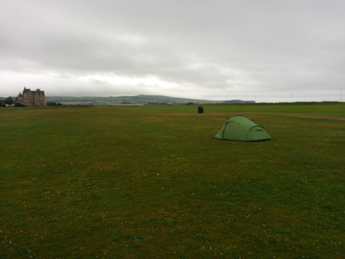Small, olive-green, dome-shaped tent pitched on a sprawling expanse of short, slightly unkempt grass. The tent sits slightly off-center, positioned in the middle ground of the image. No people are visible. In the far background, a large, imposing stone building, resembling a castle or stately home, stands subtly against the horizon. The scene is a wide, open field, seemingly coastal or near a shoreline, based on the flatness of the land and the distant hint of water or a low-lying area on the horizon. The overall colour palette is muted and cool. The grass is a deep, somewhat dull green, suggesting late summer or early autumn. The sky is a uniformly overcast, pale grey, devoid of strong light and shadow. The distant building is a light grey-brown, blending somewhat with the background. The overall lighting is soft and diffuse, lacking strong contrasts. The colour saturation is low, contributing to a subdued and somewhat melancholic mood.