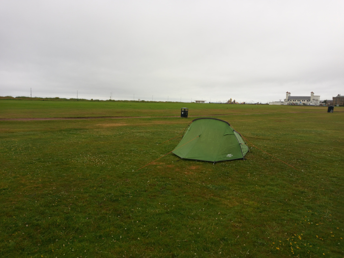 Wide, slightly low-angle shot of a solitary, lime-green, dome-shaped tent pitched on a short-cropped, verdant lawn. The tent is the central focus, occupying a significant portion of the mid-ground. Its vibrant green hue contrasts sharply with the muted tones of the surrounding landscape. It's a single-person or small two-person tent, compact and seemingly lightweight. The tent's fabric shows slight wrinkles and folds indicative of recent setup. Subtle orange guy lines are barely visible, extending from its corners to anchor it to the ground. The brand logo is faintly discernible on the tent's side. The lawn is a uniform, short-cut expanse of grass, predominantly a deep, slightly muted green. Small, scattered yellow wildflowers pepper the grass, adding subtle textural and colour variations. The grass extends to the horizon, creating a vast, flat plane. A faint, barely visible path cuts across the lawn, somewhat near the tent's placement.