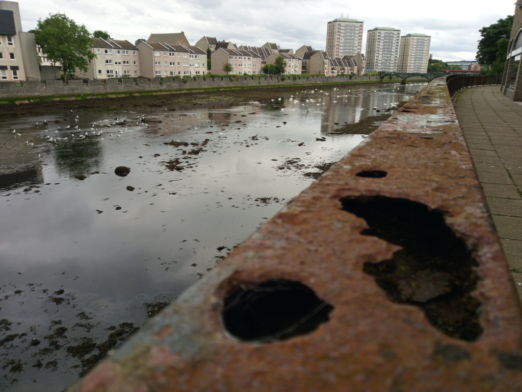 Heavily rusted, thick metal beam or barrier, positioned diagonally across the lower two-thirds of the frame. Large, irregular holes pockmark its surface, revealing glimpses of dark earth and vegetation growing within the rust. The texture is rough, uneven, and shows significant deterioration. In the background, numerous seagulls are scattered across a calm, dark-grey-green waterway. They appear mostly stationary, with a few seemingly in motion. The waterway is relatively still, reflecting the cloudy sky and the buildings on the opposite bank. The water itself has a muted palette of dark greens and greys, punctuated by patches of exposed mud or rocks. The background features a row of low-rise apartment buildings in muted beige and cream tones, stretching across the entire horizon. Behind the buildings, several taller, multi-story apartment blocks stand out, their pale concrete facades contrasting with the overcast sky. The sky is a uniform, light grey, indicative of an overcast day. The lighting is soft and diffused, devoid of harsh shadows. The colors are predominantly muted and desaturated, creating an overall feeling of calmness and even melancholy. A paved walkway appears on the right, running alongside the rusted beam, extending into the far background.