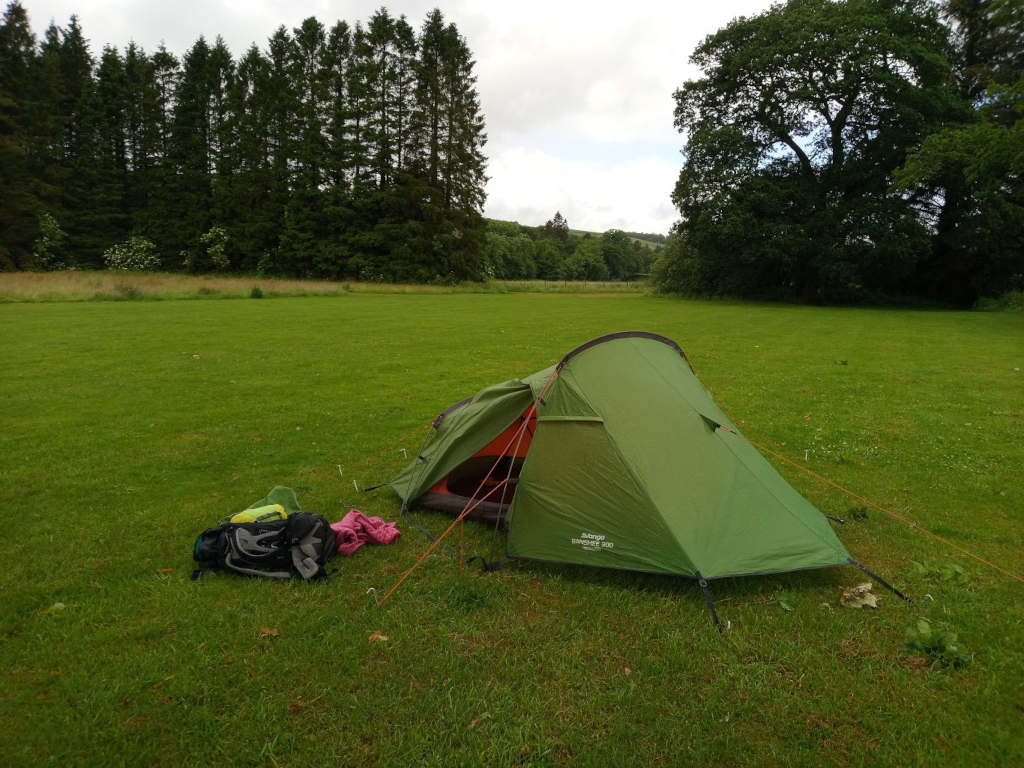 Vibrant green, single-person dome tent, pitched on a lush green lawn. It’s a compact tent, with the recognisable logo of a well-known outdoor brand partially visible near the base.  The tent is slightly angled, suggesting a natural placement rather than a meticulously straight setup. Near the tent’s base lie a backpack, a lime green sleeping bag (partially visible) and a darker pink/fuchsia colored item which could be clothing or a towel. The orange tent pegs are visible, scattered near the tent, indicating a recent setup. The ground is short-cropped grass of a consistent deep green.