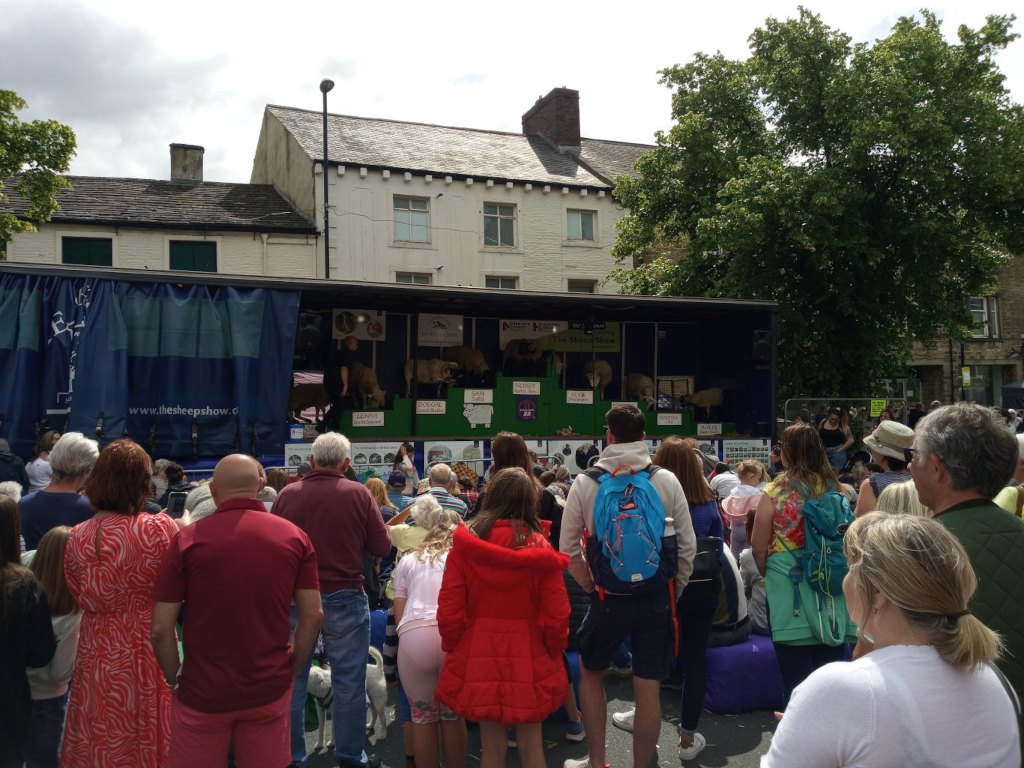 Large, flatbed truck displaying a sheep-showing event. The truck's sides are adorned with a navy blue tarp featuring the website www.thesheepshow.co.uk and images relating to sheep breeds. Inside the truck, several sheep of different breeds are visible within individual pens, with signage identifying each breed. A person in a dark shirt is seen tending to the sheep. The truck is surrounded by a large crowd of people, mostly adults and children.  Many are standing, some seated on the ground. The crowd consists of people of various ages and ethnicities, dressed in casual summer attire—bright, colourful clothing is prominent. A few people are holding small children. Notice the women in vibrant, patterned summer dresses; one is notably wearing a striking red jacket. Many people are facing towards the truck, seemingly engaged in watching the sheep presentation. Some individuals in the background are less clearly visible but still contribute to the overall density of the crowd.