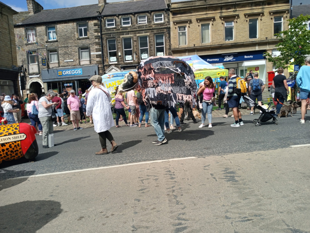 Large, whimsical sheep puppet, predominantly grey and black with textured fringe, being carried by a person mostly obscured by the puppet's bulk. The puppet's head is oversized and features faux wool and dark eyes. The carrier is dressed in dark jeans and a dark top. Surrounding this focal point is a bustling crowd of people, mostly casually dressed in summer attire. Many are observing the puppet, while others engage in seemingly everyday activities—walking, chatting, pushing strollers. A woman in a long white coat stands slightly off-centre, to the left of the puppet. She is wearing a hat, and appears to be observing the scene. Several people are visibly interacting—a group appears to be talking together, while others seem to be part of families. Noteworthy is the person in pink, who stands slightly apart from the crowd, allowing for a visual break near the puppet. Two dogs are visible in the background, one light-coloured and one darker. In the bottom left corner, a portion of a brightly coloured, patterned object is seen, possibly a vehicle or float.