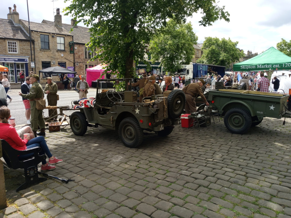 Two vintage military vehicles, a Jeep and a smaller, flatbed truck, parked on a cobblestone street. A US army olive-green Jeep, showing signs of age but seemingly well-maintained, is positioned slightly off-center, angled to the left. A small American flag is draped across the front. Its tires are large and rugged. A smaller, olive-green military-style truck, also showing its age, is parked behind and slightly to the right of the Jeep. It’s a flatbed with some equipment on the back. Several people in period attire, suggestive of World War II re-enactors, are standing near the vehicles. They are dressed in olive drab uniforms, some in officer-like attire. One man in a uniform is actively engaged with something near the back of the truck. A young woman in contemporary clothing, wearing a red shirt, sits on a park bench on the lower left, seemingly watching the scene with a companion who sits next to her. More people, in both contemporary and period-appropriate clothing, are visible in the background, suggesting a fair or market event.