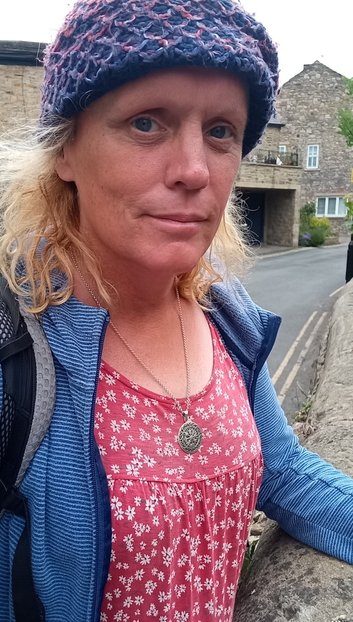 Middle-aged woman (Leonie) with shoulder-length, slightly wavy, light blonde hair. She's wearing a vibrant purplish-blue hand-knitted beanie, pulled down to her eyebrows, which partially obscures her forehead. Her skin is fair with a slightly sun-kissed tone, showing some fine lines and texture, indicative of age and time spent outdoors. Her expression is neutral, bordering on slightly pensive; her gaze is directed slightly to the camera's left. She wears a blue ribbed knit jacket over a coral-red dress speckled with small white flowers. The dress has a gathered bodice and a simple, slightly A-line skirt. A silver chain necklace featuring a circular, slightly ornate pendant hangs just below her neckline. She leans slightly against a stone wall, her left arm resting on the wall, while her right arm hangs loosely by her side. A portion of a gray backpack is visible on her left shoulder. The background is slightly out of focus but reveals a quaint stone street scene. A stone building with multi-paned windows and a muted gray-brown colour palette is visible behind her. The sky is overcast, producing soft, diffused daylight that avoids harsh shadows.