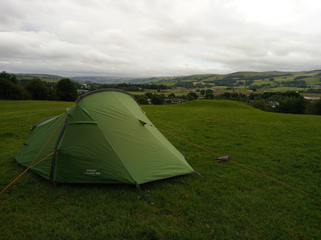 Two-person, olive-green tent, a Vango Banshee 300, pitched on a lush, short-cropped grassy field. The tent is positioned slightly off-centre, angled towards the viewer's left, revealing its partially open doorway. Its guy lines, a vibrant burnt orange, extend from the tent's corners, anchored to the ground. A small, dark gray object, possibly a tent stake bag or a small rock, lies near one of the guy lines. The tent appears new or well-maintained, exhibiting a clean, smooth texture. The scene is set in a rolling, pastoral landscape under a soft, overcast sky. The sky is a muted greyish-white, with no harsh sunlight. The background displays a series of gently rising hills and valleys, dotted with patches of green fields, trees, and hints of distant buildings or structures suggesting a small town or village nestling in the valley.