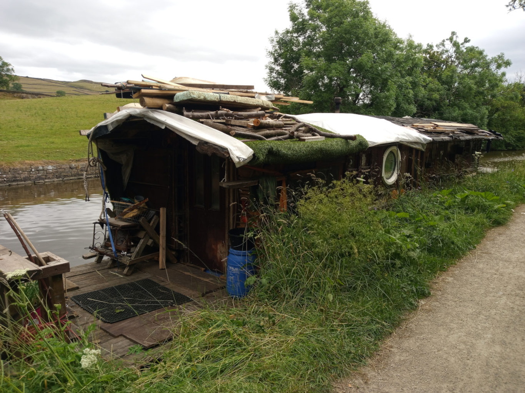 Dilapidated, narrowboat-like structure moored on a canal. Its wooden hull is weathered and dark brown, showing significant age and wear. The roof is partially covered by a tattered white tarp, under which branches, logs, and various pieces of lumber are haphazardly piled, creating a wild, overgrown look.  Patches of moss and grass grow on the roof and sides of the boat. A porthole, circular and framed in dark wood, is visible near the centre of the structure's side.