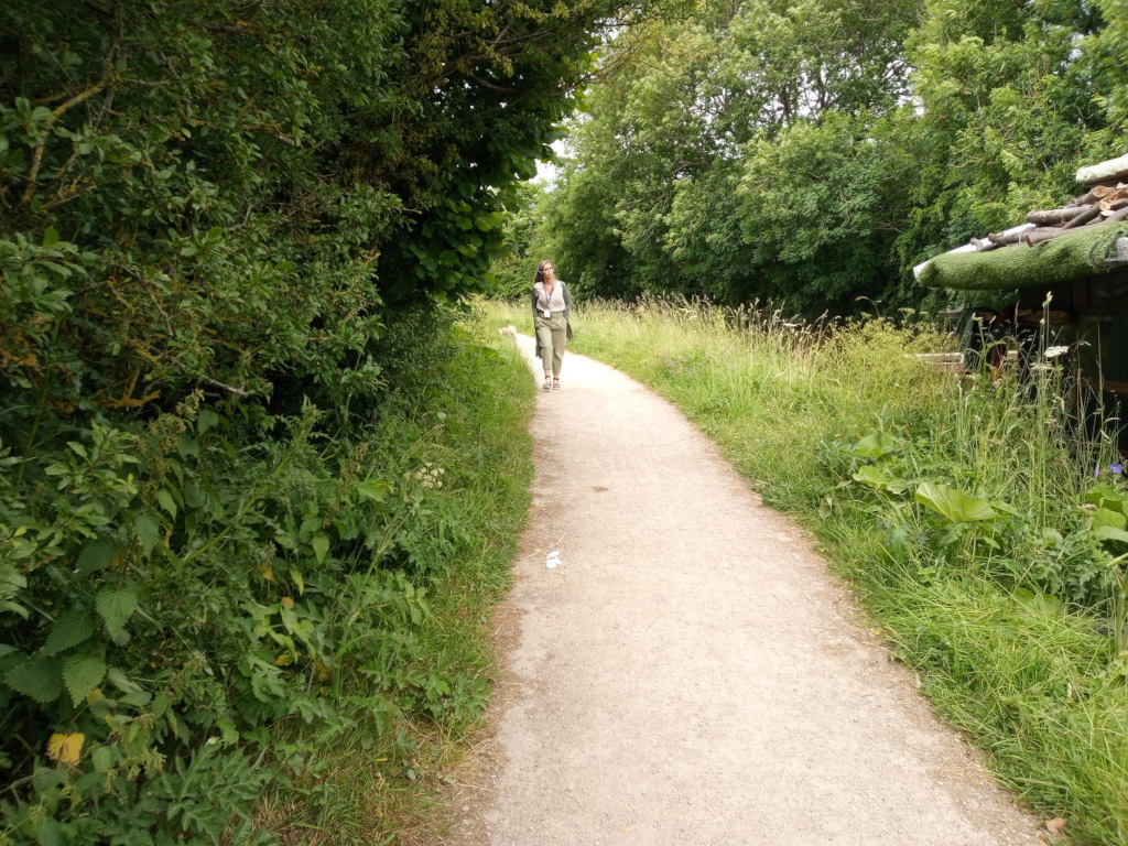 Woman walking away from the viewer down a gravel path. She's positioned slightly off-centre, towards the left, and appears to be of medium build. She's wearing light beige or khaki-coloured pants and a similarly light-coloured, possibly linen or cotton, loose-fitting shirt or blouse. Her hair is light-brown and seems to be pulled back from her face. She’s walking at a relaxed pace, not hurried. In the background to the right, a partially visible rustic structure is present; it seems to be a small, simple building with a thatched or green-covered roof, hinting at a possibly rural or historical setting. The structure is partially obscured by tall grasses.