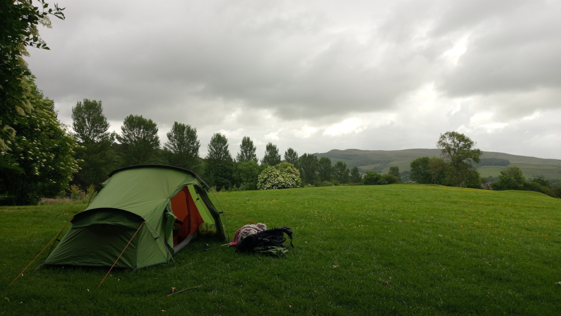 A green two-person tent pitched on a grassy field. A backpack lies next to the tent. In the background, there are trees and rolling hills under a cloudy sky. The overall mood is serene and peaceful, suggesting a camping trip in nature. The weather appears overcast.