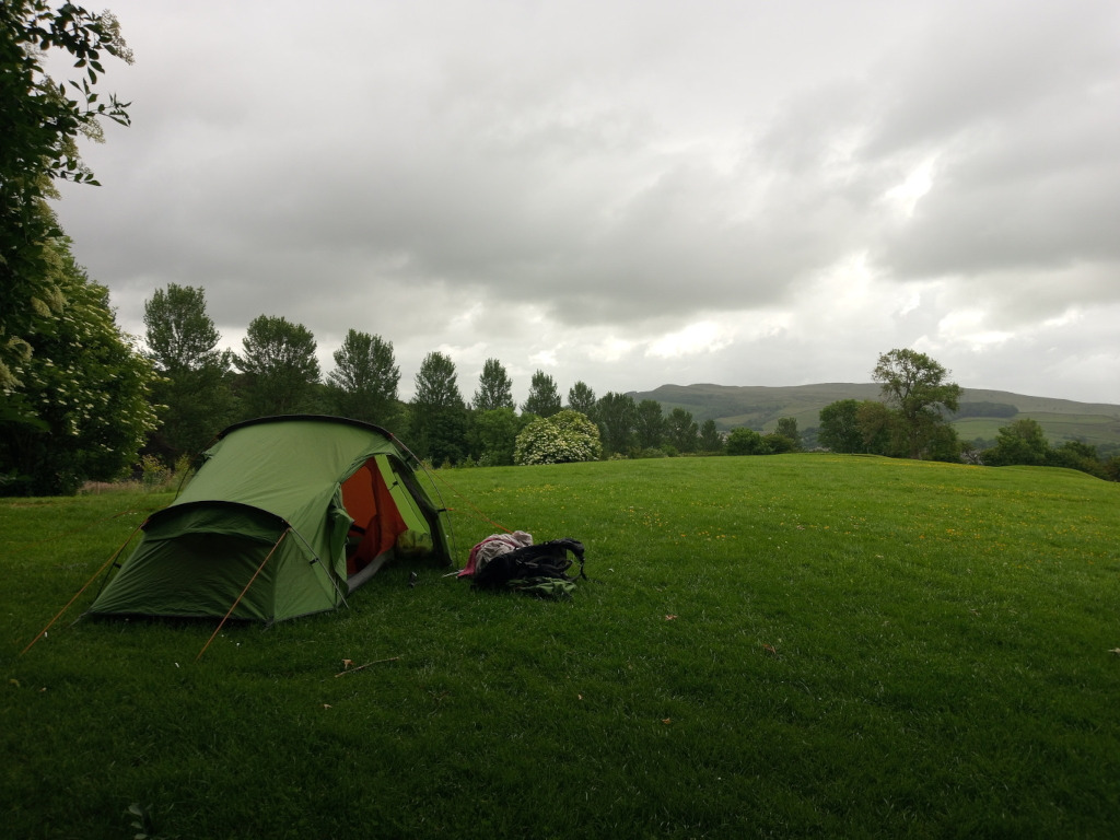 Green two-person tent pitched on a grassy field. A backpack lies next to the tent. In the background, there are trees and rolling hills under a cloudy sky. The overall mood is serene and peaceful, suggesting a camping trip in nature. The weather appears overcast.