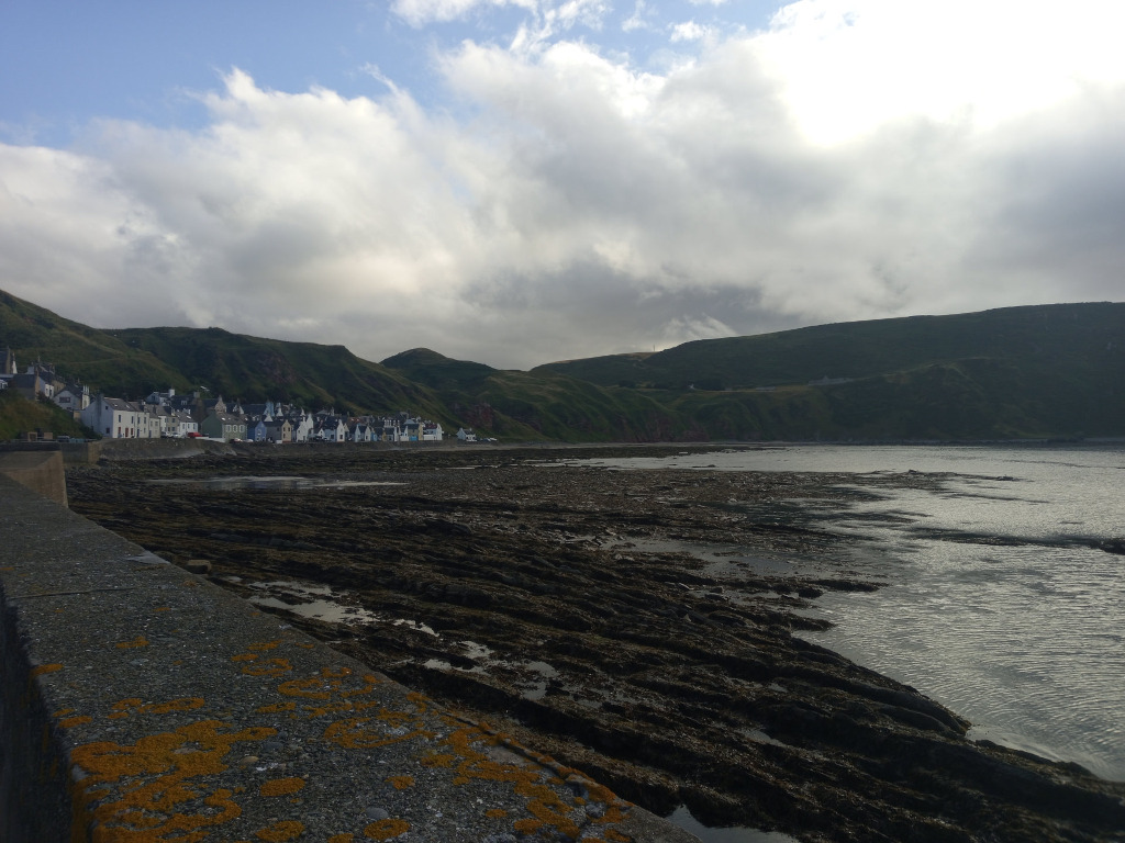 Coastal scene, likely in a small village. The foreground features a low, moss-covered wall next to a rocky inter-tidal zone. In the mid-ground, a row of small, whitewashed houses lines the shore at the base of a green hill or cliff. The background is dominated by a larger, darker green hill under a partly cloudy sky. The overall impression is of a serene, somewhat secluded, coastal setting.