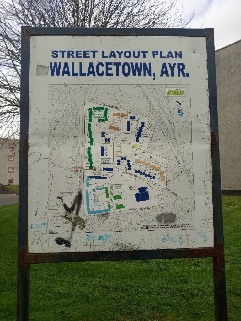 Weathered street layout plan sign for Wallacetown, Ayr. The sign displays a map of the area, indicating streets, buildings, and possibly blocks of flats. The map is somewhat faded and has graffiti on it. The sign itself shows signs of age and exposure to the elements.
