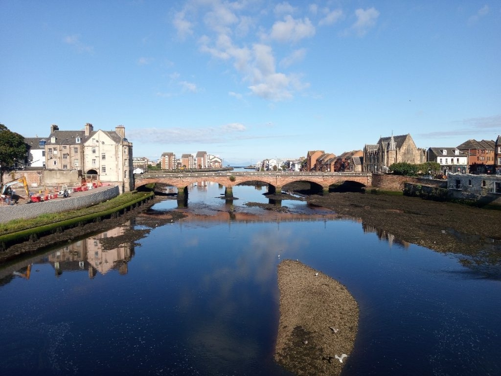 River scene, likely in a European town. A stone bridge arches elegantly across the river, connecting two sides lined with historical buildings, some reflecting in the calm waters. A small, rocky islet sits in the middle of the river, adding a touch of natural intrigue. The sky is clear and bright, enhancing the overall peaceful atmosphere of the scene. Construction equipment is visible on the left bank, indicating ongoing development or renovation. The architecture suggests a blend of historical and modern buildings, hinting at a town rich in history but also experiencing growth and change.