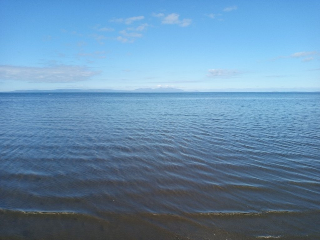 Tranquil seascape. The foreground shows the gently lapping waves on a sandy beach. The middle ground is a calm expanse of sea, its surface marked by small ripples. In the far distance, a low-lying landmass or distant shore is visible against a clear, blue sky with a few wispy clouds.