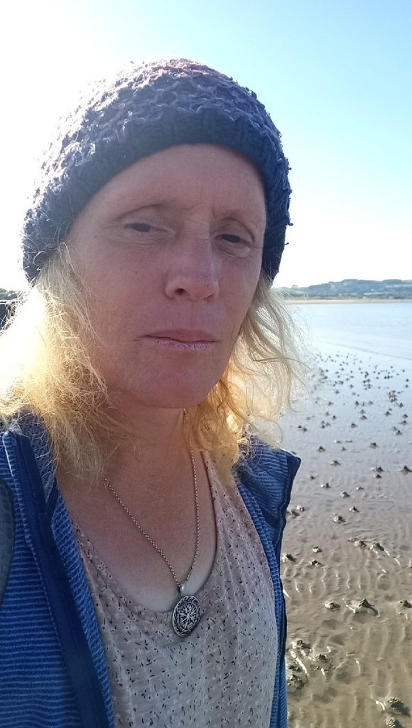 Blonde woman wearing a blue knitted hat and a blue jacket. She is wearing a beige top and a necklace with a circular pendant. Her expression is serious or pensive. The background is a beach or mudflat with many small holes or indentations, and a calm body of water and distant land in the far background.