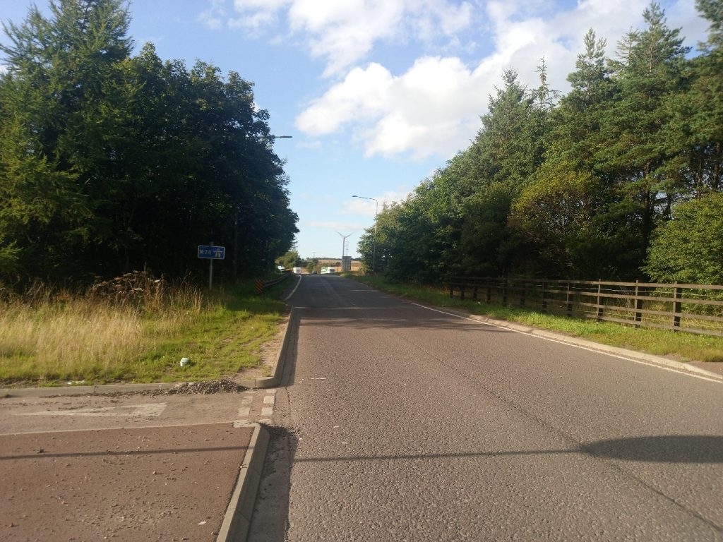 View down a relatively straight, two-lane road. The road is bordered on both sides by lush green trees and vegetation. On the left, there's a small grassy area with some taller weeds, and a roadside sign indicating M74 is visible. A wooden fence runs along the right-hand side of the road. In the distance, a wind turbine can be seen under a bright, partly cloudy sky.