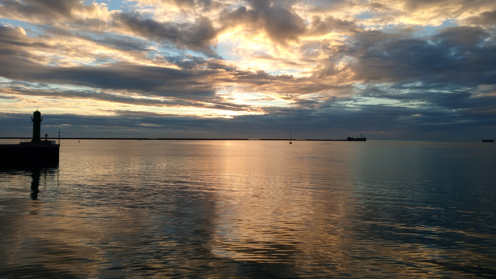 A tranquil coastal scene at sunset. A small lighthouse stands on a pier to the left, silhouetted against a sky filled with dramatic, colorful clouds.  The sun is low on the horizon, casting a golden glow on the calm water, which reflects the sunset hues.  In the distance, a small sailboat and other structures are barely visible. The overall mood is serene and peaceful, showcasing the beauty of a natural sunset over the sea.