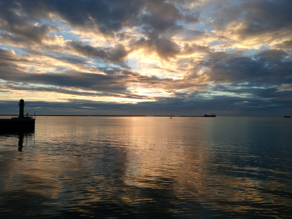 Tranquil coastal scene at sunset. A small lighthouse stands on a pier to the left, silhouetted against a sky filled with dramatic, colourful clouds.  The sun is low on the horizon, casting a golden glow on the calm water, which reflects the sunset hues.  In the distance, a small sailboat and other structures are barely visible. The overall mood is serene and peaceful, showcasing the beauty of a natural sunset over the sea.