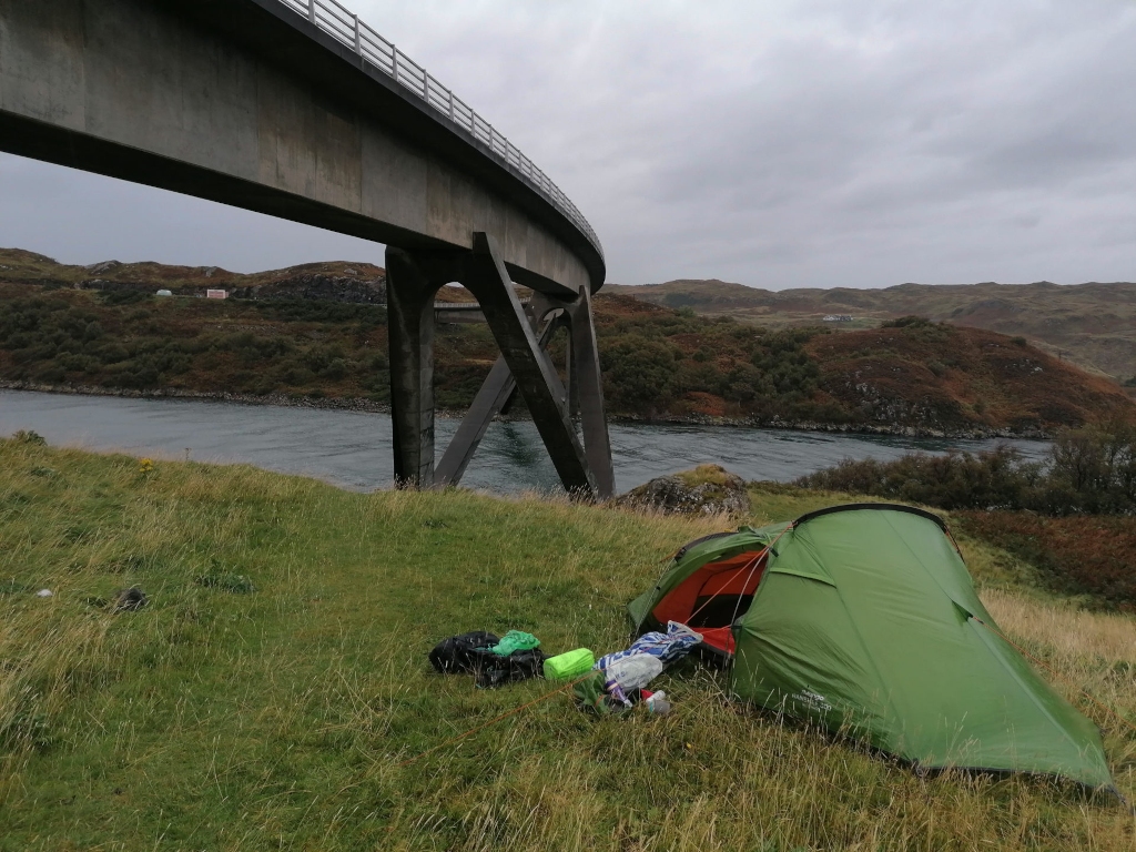 Vibrant green, single-person dome tent pitched on a grassy hillside. The tent is slightly angled, its entrance facing the viewer. Around the base of the tent lie a few items of camping gear: a dark-coloured backpack or duffel bag, a bright lime green bag (possibly a dry bag or stuff sack), and other indistinct smaller items. There's no person visible. The scene is set in a dramatic, rugged landscape. The hillside is covered in lush, short, slightly overgrown green grass, typical of a coastal region. In the background, a large concrete bridge sweeps across a calm, dark-grey-blue body of water, stretching from the lower left to the middle right of the frame, curving gently. Beyond the water, there are rolling green and brown hills under a pale grey, overcast sky.