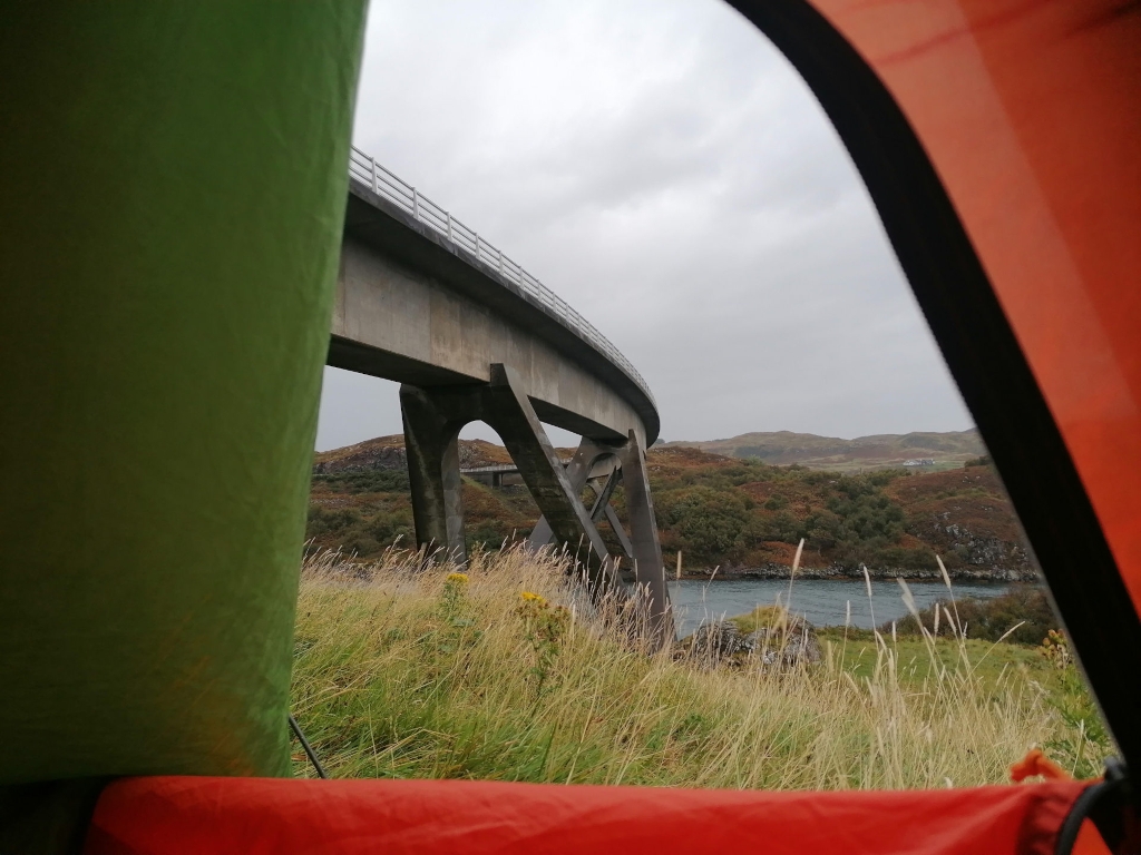 Interior of a two-toned tent. A vibrant orange section occupies the bottom and right, while a muted olive-green section takes up the left and upper portion. The tent fabric is slightly wrinkled and textured, visible only from the inside looking out. The tent’s edges frame the scene, acting as a natural border. The grass immediately outside the tent is tall, dry-looking, and golden-brown in patches, with hints of late-summer wildflowers.