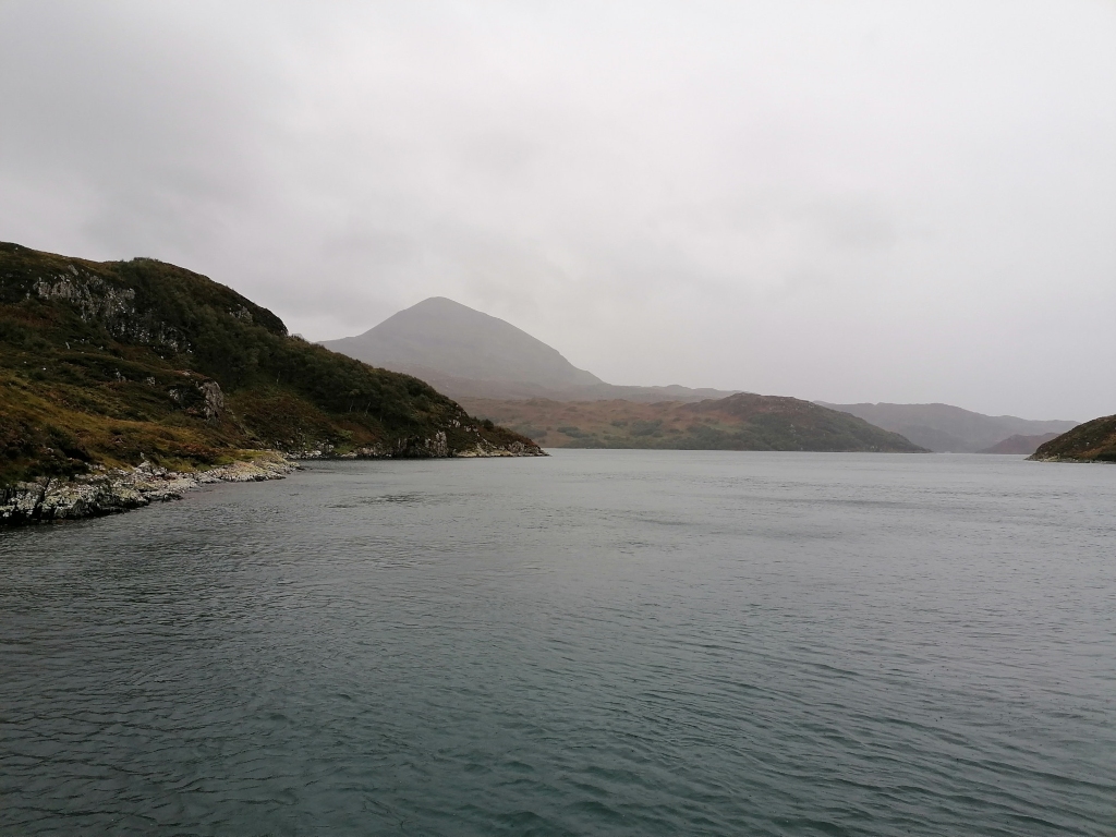 Serene, expansive landscape dominated by a body of water, likely a loch or fjord. The foreground is occupied by the calm, dark-grey-blue water, its surface subtly rippled. The mid-ground shows a narrow waterway, separating two landmasses.  In the background, rolling hills and mountains form a rugged coastline. No people or other animals are visible. The scene is set under an overcast sky, a uniformly pale grey with no visible sun. The light is diffuse and soft, lacking harsh shadows, contributing to a muted, somewhat melancholic ambiance. The color palette is predominantly muted earth tones: dark greens on the vegetation-covered hills, the grey-blue of the water, and the pale grey of the sky. The mountains in the distance are a blend of muted greens, browns, and greys.