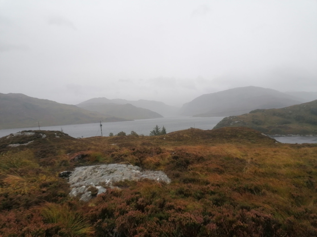 Landscape featuring a gently sloping, heather-covered hill in the foreground. This hill is predominantly composed of muted browns and russets, interspersed with patches of dull, grayish-green grass. A large, light gray boulder, weathered and irregular in shape, sits prominently in the mid-ground of the foreground hill. In the far background, there are two hazy, indistinct telephone poles standing near the water's edge. The scene is set in a remote, mountainous region, likely coastal, given the presence of a body of calm, dark gray-blue water. The water, nestled between imposing, hazy mountain ranges, stretches from the mid-ground to the horizon. These mountains appear soft and indistinct due to a heavy mist or light rain, their shapes barely defined by the diffuse light. The overall colour palette is subdued and muted, characterised by various shades of grey, brown, and green, creating a monochromatic feel dominated by cool tones. The lighting is diffuse and even, lacking sharp shadows or strong highlights due to the overcast sky. The atmosphere is damp and cool, suggested by the misty air.